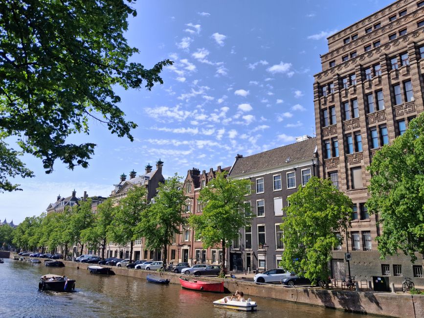 Capture of a scenic canal in Amsterdam with historic buildings and lush trees under a clear summer sky.