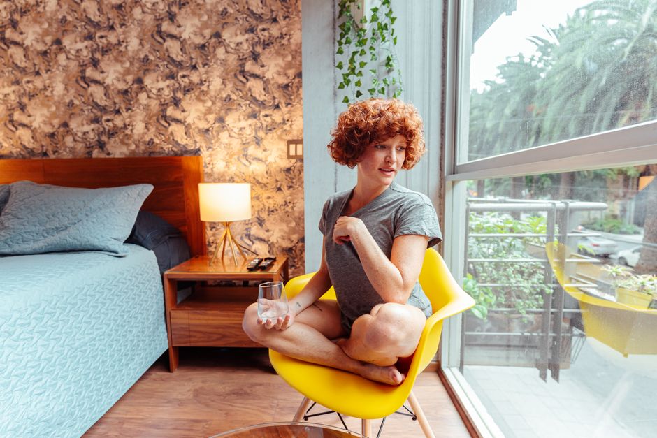 Woman with curly hair sitting on a chair by a window, holding a glass in a cozy bedroom.