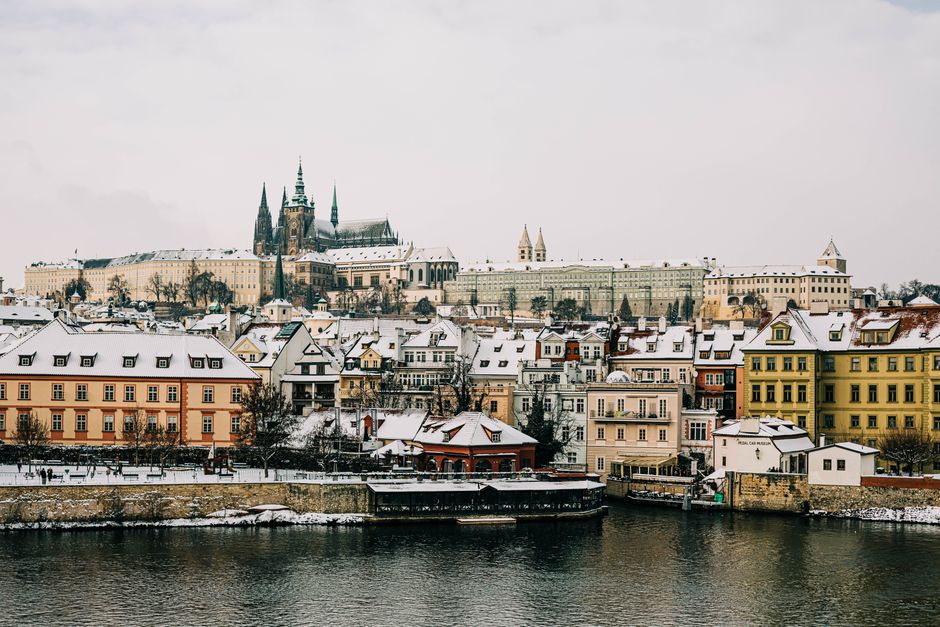 Explore a stunning winter view of Prague with snow-covered rooftops and historic architecture along the river.