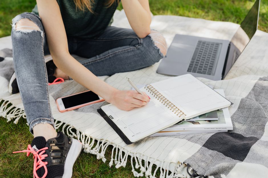 Crop faceless female student in casual clothes taking notes in planner while sitting on blanket with smartphone and laptop on green park grass on sunny day