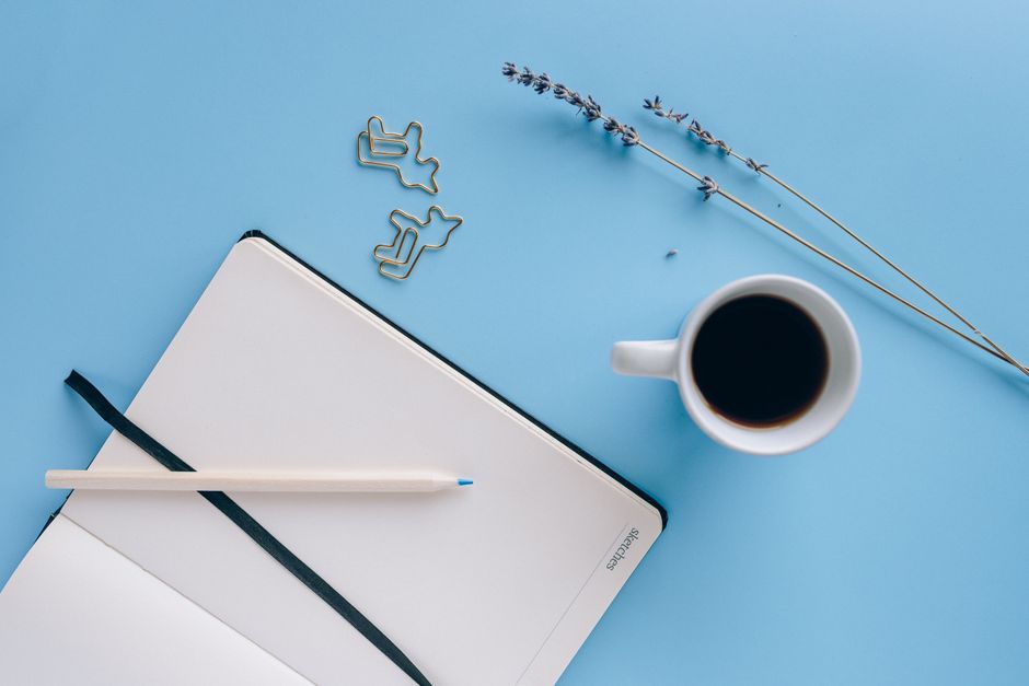 Flatlay of a minimalist workspace featuring a mug of coffee, open notebook, and lavender on a blue background.
