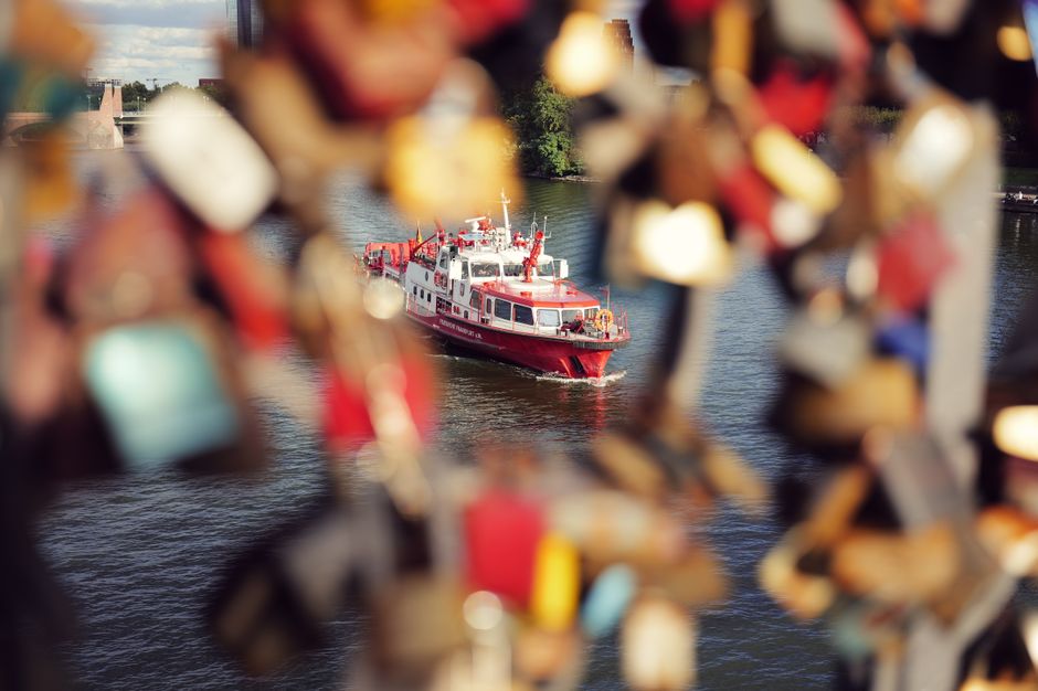 Red boat cruises on the Main River in Frankfurt, framed by vibrant love locks on a bridge.