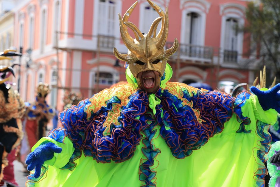 A performer in colorful costume and mask at a lively outdoor carnival parade.